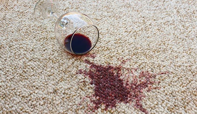 A clear glass of red wine is lying on its side on a light-colored rug with a dark red wine stain spilling out